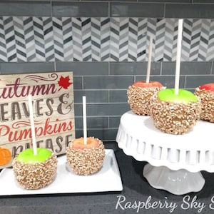 May include: Four caramel apples decorated with chopped nuts on white sticks. The apples are on a white cake stand and a white rectangular plate. A wooden sign with the text "Autumn Leaves & Pumpkins Please" is in the background.