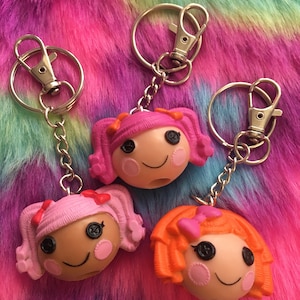 May include: Three doll-faced keychains with pink, light pink, and orange hair. Each keychain has a silver clasp and chain. The dolls have button eyes, rosy cheeks, and smiling faces. The background is a colorful, fluffy texture.