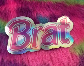 May include: A holographic sticker with the word "Brat" in pink, outlined in a lighter pink, and a silver border. The sticker is on a vibrant, multi-colored, faux fur background.