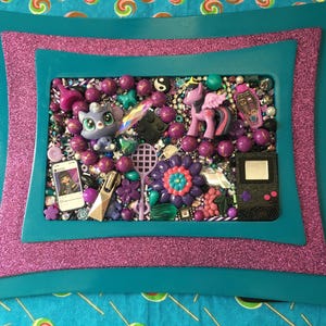 May include: A rectangular frame with a teal border and a purple glitter inner layer containing a collage of purple and teal beads, a small purple pony figure, a black handheld game console, a miniature smartphone, and a purple flower ornament.