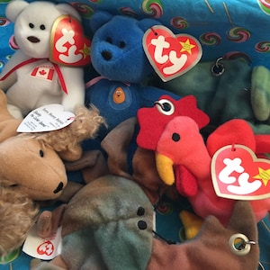 May include: A collection of colorful Ty Beanie Babies plush toys, including a white bear with a Canadian flag, a blue bear, a brown dog, a green caterpillar, and a red rooster. Each toy has a heart-shaped tag.