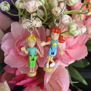 May include: A pair of dangle earrings featuring miniature doll figures. One doll has blonde hair, a teal top, and green pants. The other doll has red hair and a blue dress. Both dolls are on small, round bases and are suspended from silver-tone ear wires.
