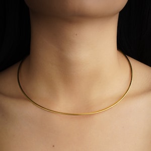 Op de afbeelding: Een gouden chokerketting om de nek van een persoon. De ketting is een eenvoudige, dunne metalen band.