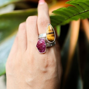 May include: Two silver rings with gemstone settings. The ring on the bottom features a red ruby, while the ring on top features a teardrop-shaped tiger's eye stone.