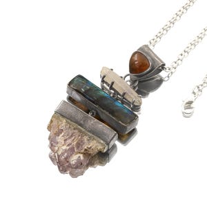 May include: A silver necklace with a pendant composed of various gemstones. The pendant incorporates a rough amethyst, a rectangular labradorite, a clear quartz crystal, and a small, triangular, orange-brown stone. The necklace chain is silver.