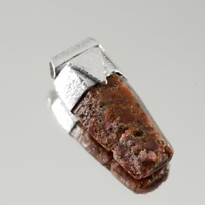 May include: A raw, reddish-brown gemstone pendant with a silver setting. The gemstone is rough and has a natural, unpolished look.