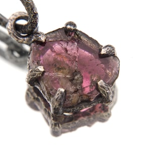 May include: A close-up of a pendant featuring a raw, pink gemstone set in a textured, dark metal. The gemstone has a rough, natural appearance, and the metal setting has a detailed, organic design. The pendant hangs from a matching metal loop.