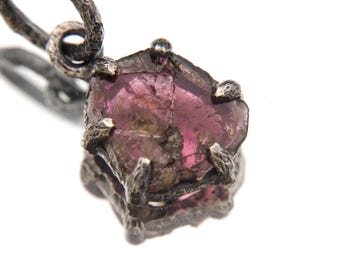Puede incluir: Primer plano de un colgante con una gema rosa en bruto engastada en un metal oscuro texturizado. La gema tiene un aspecto rugoso y natural, y el engaste metálico presenta un diseño orgánico detallado. El colgante cuelga de un lazo de metal a juego.