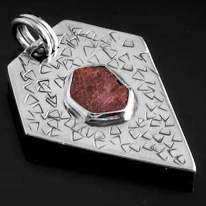 Puede incluir: Un colgante de plata con un diseño geométrico y una piedra preciosa roja en el centro.