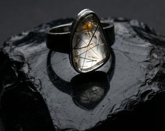 May include: A silver ring with a clear, triangular gemstone. The gemstone has black and gold inclusions, set against a dark background. The ring is a unique piece of jewelry.