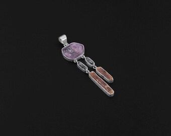 May include: A silver pendant with a large, light purple, irregularly shaped stone at the top. Two smaller, rectangular stones in shades of brown and gray hang below. The pendant is set against a black background.