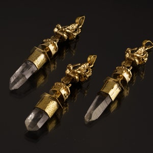May include: Three gold pendants with clear quartz crystals and intricate gold designs. The pendants are on a black background.