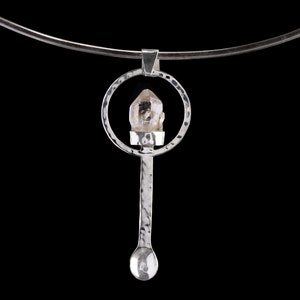 Könnte beinhalten: Eine silberne Halskette mit einem Anhänger, der einen klaren Quarzkristall in einem Kreis umschließt. Der Anhänger hängt an einer gehämmerten Silberkette.