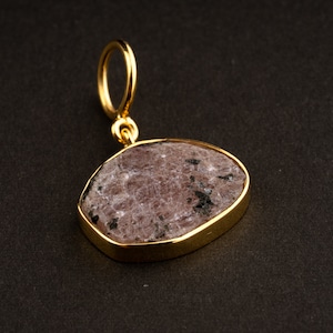 Puede incluir: Un colgante de oro con una piedra preciosa de color púrpura claro y negro. El colgante tiene forma ovalada y un acabado liso y pulido.