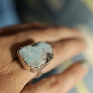 以下が含まれることがあります： 大きな、不規則な形の宝石をあしらったシルバーリング。石は明るい青色で、より暗いインクルージョンがあります。リングはシンプルで磨かれたシルバーバンドにセットされています。宝石のユニークな質感と色に焦点が当てられています。