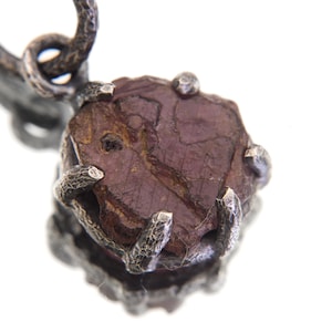 Puede incluir: Primer plano de un colgante con una piedra preciosa en bruto, de color granate, sujeta en una montura de plata. La plata tiene un acabado texturizado y oxidado, y la piedra preciosa tiene forma irregular. El colgante está suspendido de un lazo de plata.
