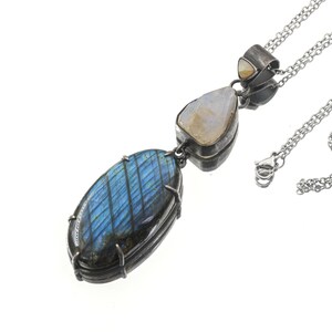 Puede incluir: Un collar de plata con tres colgantes de piedras preciosas. El más grande es un labradorita ovalada con iridiscencia azul y verde. Los otros dos son más pequeños, con una piedra lunar blanca nacarada y una piedra pequeña de color claro.