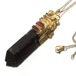 Puede incluir: Un collar dorado con un colgante de cristal marrón oscuro con punta facetada. El colgante está adornado con metal dorado y dos pequeñas piedras preciosas rosas rectangulares. El collar tiene una cadena delicada.