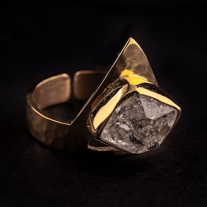 Puede incluir: Un anillo dorado con una piedra transparente grande e irregular. El anillo tiene un diseño único con una banda abierta y un engaste geométrico para la piedra. El anillo está sobre un fondo negro.