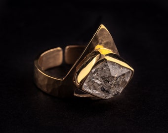 May include: A gold-colored ring with a large, irregularly shaped, clear stone. The ring has a unique design with an open band and a geometric setting for the stone. The ring is set against a black background.