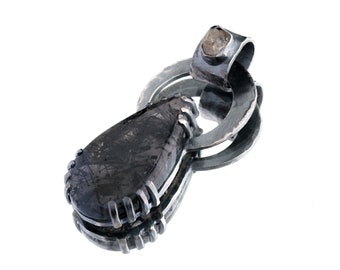 Puede incluir: Un colgante en forma de lágrima con una piedra oscura y texturizada con inclusiones visibles, engastada en una montura de plata con borde dentado. El colgante incluye un aro circular de plata en la parte superior con un pequeño acento de piedra.
