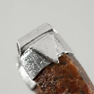 May include: Close-up of a silver pendant with a rough, brown stone. The silver is textured and has a geometric design.