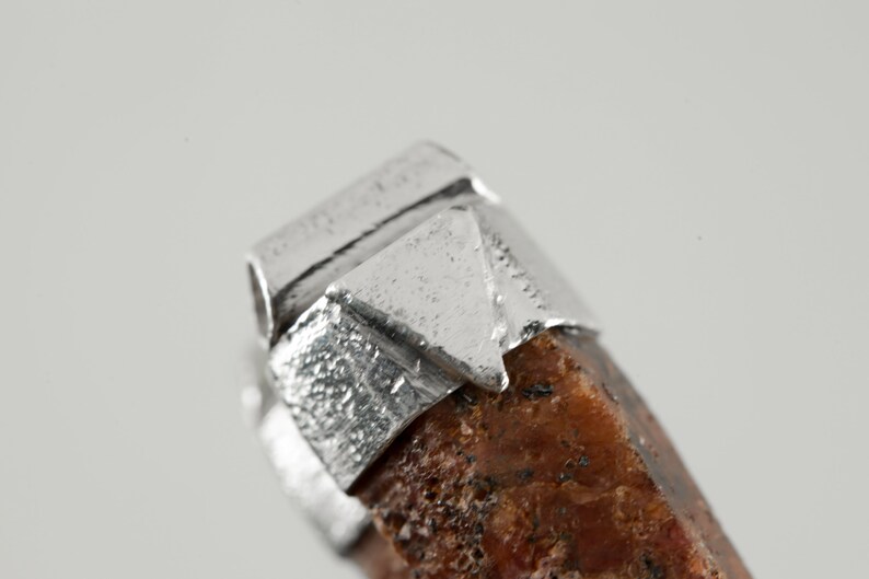 May include: Close-up of a silver pendant with a rough, brown stone. The silver is textured and has a geometric design.