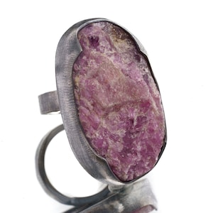 May include: A silver ring featuring a large, oval-shaped, pink gemstone set in the centre. The gemstone has a rough, textured surface.