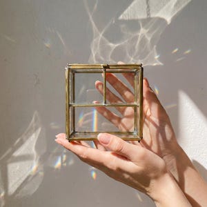 May include: A small, square glass and brass box with a hinged lid. The box is held in open hands, with sunlight creating a prism effect on the wall behind it. The box has a decorative, vintage aesthetic.