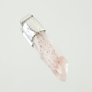 May include: A clear quartz crystal pendant with a silver setting. The crystal has a pink hue and is speckled with small red inclusions.