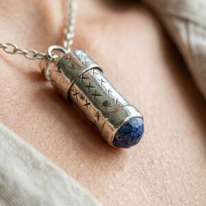 May include: Silver-toned pendant necklace with a cylindrical design. The pendant has etched cross-hatch patterns and a blue gemstone at the base. The necklace chain is also silver-toned.