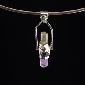Op de afbeelding: Een zilveren ketting met een hanger, met een turquoise steen aan de bovenkant, een helder kwartskristal in het midden en een paarse amethistkristal aan de onderkant.