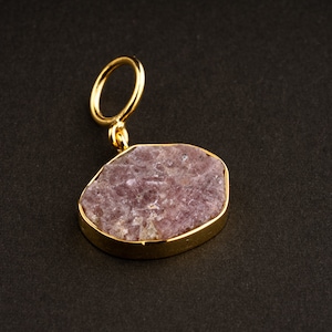 Puede incluir: Un colgante de oro con una piedra preciosa rosa y morada. La piedra preciosa tiene una forma irregular y una textura rugosa y natural.