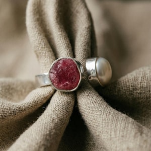 Könnte beinhalten: Zwei Silberringe mit Edelsteinen. Ein Ring hat einen großen, unregelmäßig geformten, tiefrosa Edelstein. Der andere Ring hat eine große, runde, weiße Perle. Die Ringe werden auf einem beigen Stoff präsentiert.