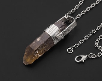 May include: A silver-toned necklace featuring a smoky quartz crystal pendant. The crystal is a light brown color with a pointed end and is encased in a textured silver setting. The chain has a lobster clasp.
