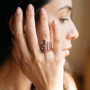 Può includere: Un anello d'argento con tre pietre rettangolari nei toni del rosso, viola e arancione. L'anello è su un dito, con la mano a fuoco. Lo sfondo è sfocato.
