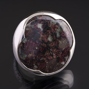 May include: A silver ring with a round, dark red and green gemstone set in a silver bezel. The ring has a unique, organic shape.
