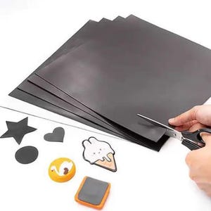 May include: A stack of black magnetic sheets with various cut-out shapes and decorative magnets. A pair of scissors is cutting the sheets. The magnets include a star, heart, circle, swan, ice cream cone, and a rectangular shape.