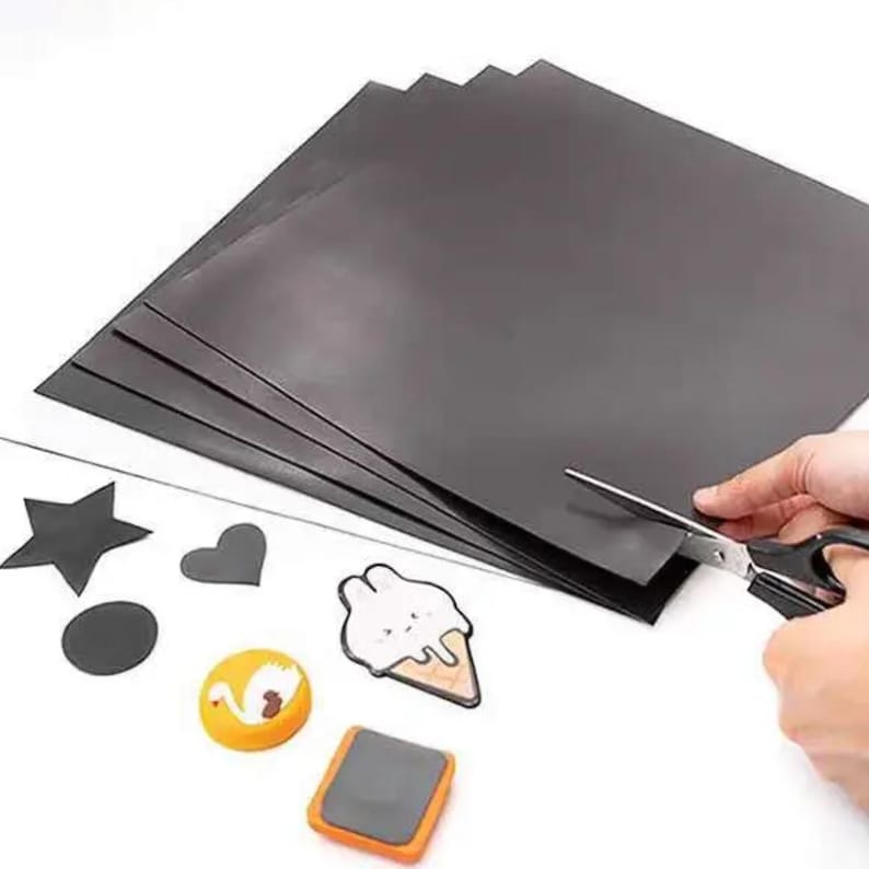 May include: A stack of black magnetic sheets with various cut-out shapes and decorative magnets. A pair of scissors is cutting the sheets. The magnets include a star, heart, circle, swan, ice cream cone, and a rectangular shape.