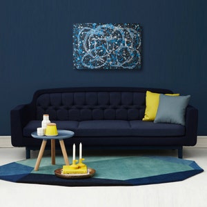 May include: A dark blue sofa with a light blue and white abstract painting hanging above it. The sofa has a yellow pillow and a gray pillow. A small wooden table with a blue top sits in front of the sofa. The table has two white ceramic containers and a yellow ceramic container on it. There are two yellow candles on the table. A blue and green geometric rug is on the floor in front of the sofa.