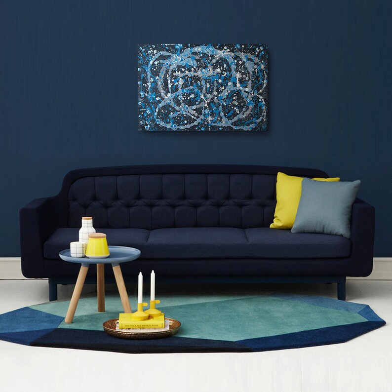 May include: A dark blue sofa with a light blue and white abstract painting hanging above it. The sofa has a yellow pillow and a gray pillow. A small wooden table with a blue top sits in front of the sofa. The table has two white ceramic containers and a yellow ceramic container on it. There are two yellow candles on the table. A blue and green geometric rug is on the floor in front of the sofa.