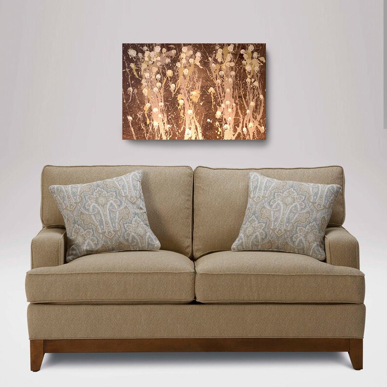 May include: A beige sofa with two pillows featuring a paisley pattern. The sofa is in front of a white wall with a brown abstract painting above it.
