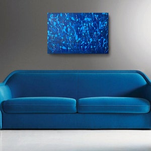 May include: A blue velvet sofa with a modern design sits in front of a gray wall with an abstract blue and white painting hanging above it.