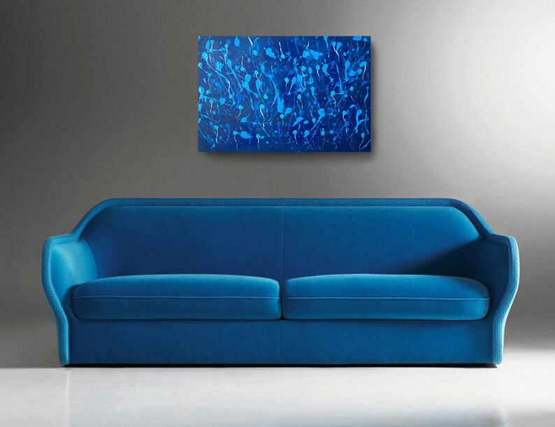 May include: A blue velvet sofa with a modern design sits in front of a gray wall with an abstract blue and white painting hanging above it.