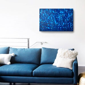 May include: Abstract painting with a blue background and white splatter paint. The painting is hanging on a white wall above a blue couch.