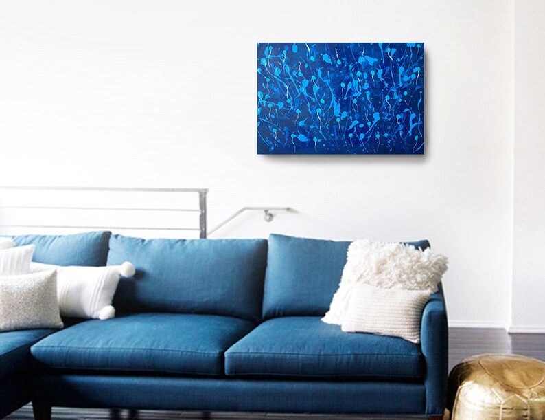 May include: Abstract painting with a blue background and white splatter paint. The painting is hanging on a white wall above a blue couch.