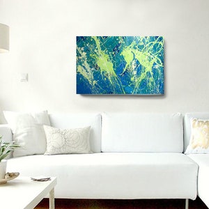 May include: Abstract art canvas with a vibrant display of blue, green, and white paint splatters. The artwork is mounted on a white wall above a white sectional sofa. The room is decorated with a white lamp, vase, and pillows.