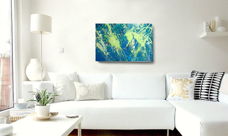 May include: Abstract art canvas with a vibrant display of blue, green, and white paint splatters. The artwork is mounted on a white wall above a white sectional sofa. The room is decorated with a white lamp, vase, and pillows.