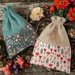 May include: Two drawstring bags with floral patterns and the letter "A" embroidered on them. One bag is teal with a floral design, and the other is beige with a floral and heart pattern. Both bags have a natural twine drawstring.