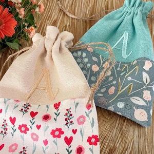 May include: Two drawstring bags with floral patterns and embroidered letters. One bag is light beige with a floral pattern of red flowers and hearts. The other is teal with a floral pattern and the letter "A". Both bags have a jute drawstring.